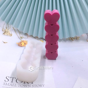 May include: A white silicone mold with four heart-shaped cavities. Next to the mold is a stack of five pink heart-shaped objects.