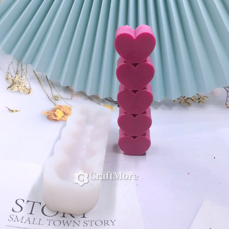 May include: A white silicone mold with four heart-shaped cavities. Next to the mold is a stack of five pink heart-shaped objects.