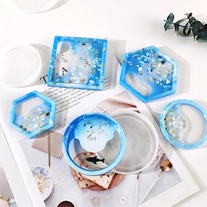 May include: Set of five blue resin coasters with a white and blue gradient and embedded seashells. The coasters are in various shapes, including square, round, and hexagonal.