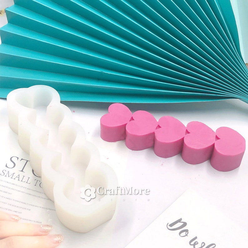 May include: White silicone mold with five heart-shaped cavities. A row of five pink heart-shaped soaps are next to the mold. The text "CraftMore" is visible on the white surface.