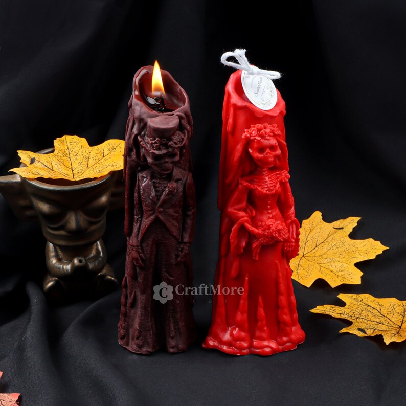 May include: Two tall, hand-painted, red and burgundy wax candles shaped like skeletons in wedding attire. The bride is wearing a red gown and the groom is wearing a burgundy suit. The candles are lit and have a white label with the text "CraftMore" on the groom's candle.