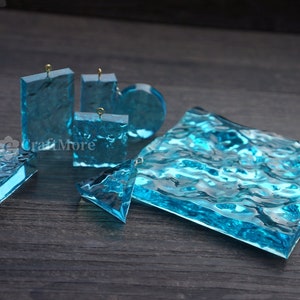 May include: A set of six clear blue resin pendants with a wave-like texture. The pendants are of various shapes and sizes, including rectangular, square, and triangular. Each pendant has a gold jump ring for attaching to a chain or cord.