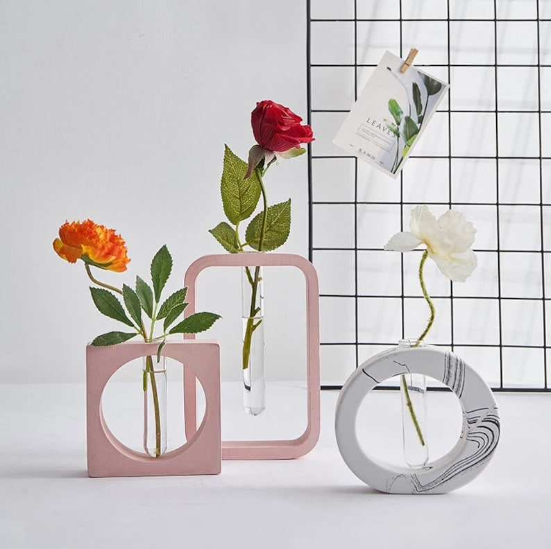 May include: Three modern geometric vases with clear glass test tubes for holding flowers. The vases are made of pink, white, and gray concrete. The vases are arranged on a white surface in front of a black wire grid.