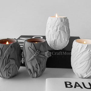 May include: Four concrete candle holders with a variety of designs. The candles are lit and the holders are on a black surface with the word "CraftMore" printed on it.