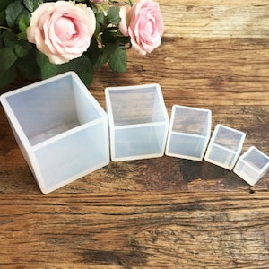 Peut inclure: Ensemble de six moules en silicone transparent de différentes tailles, en forme de cubes. Les moules sont disposés par ordre de taille, du plus grand au plus petit, sur une surface en bois.