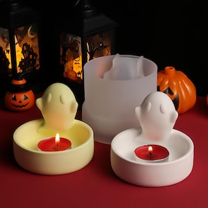 Puede incluir: Candelabros con temática de Halloween con diseño de fantasma. Uno es amarillo pálido, el otro blanco, cada uno con una vela de té roja. También se ve un molde translúcido con forma de fantasma y una pequeña calabaza naranja.