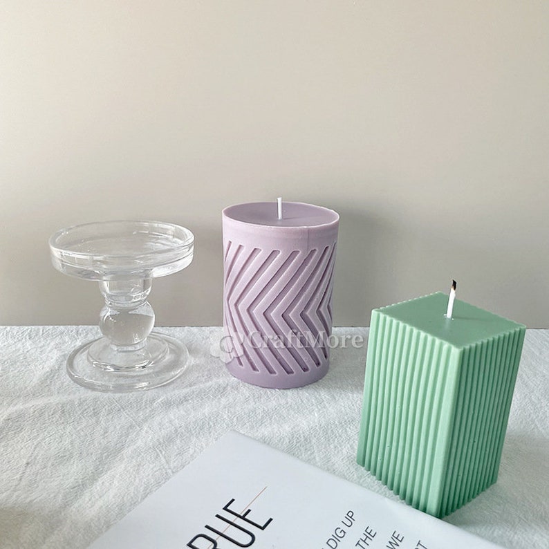 May include: Two decorative candles, one purple with a chevron pattern and one green with vertical lines, on a white surface with a glass candle holder.
