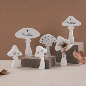Könnte beinhalten: Satz mit sechs weißen Holzpilzfiguren mit verschiedenen Designs. Einige Pilze haben Text darauf, wie "Hello sunshine" und "Tall & Story". Ein Pilz hat schwarze Punkte und ein anderer ein kleines Herz.