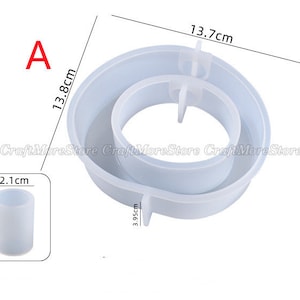 May include: A white silicone mold for making a two-tiered cake or dessert. The mold is 13.7 cm in diameter and 13.8 cm tall. It has a smaller inner ring that is 3.95 cm in diameter. The mold is shown next to a smaller silicone mold that is 2.1 cm in diameter and 2.8 cm tall.