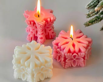 May include: Three snowflake-shaped candles. Two are lit, emitting a warm glow. The candles are pink and white, with intricate snowflake designs. The lit candles are a vibrant pink, while the unlit candle is a soft white. The candles are a festive holiday decoration.