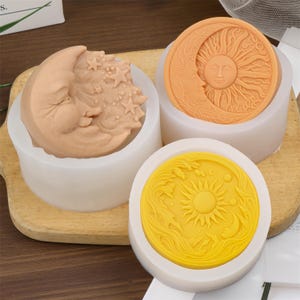 Pode incluir: Três moldes de sabão com tema celestial. Um apresenta uma lua crescente com estrelas, outro um rosto de sol e lua, e o terceiro um sol com ondas e uma lua crescente. Os sabonetes estão em moldes de silicone brancos.