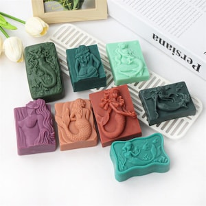 May include: An assortment of rectangular soaps in various colors, each featuring a mermaid relief design. Colors include green, purple, coral, and teal. The soaps are arranged on a white, slotted tray, with a book and decorative items in the background.