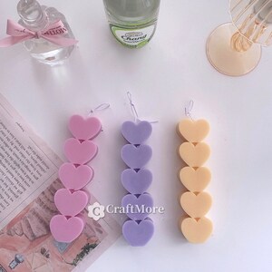 May include: Three heart-shaped candles in pink, purple, and yellow. Each candle is made up of five hearts stacked on top of each other.