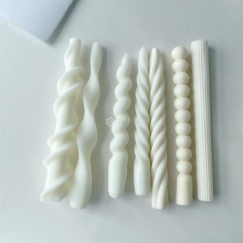 May include: A selection of white decorative candles in diverse shapes. The candles feature twisted, spiral, and column designs. The candles are displayed on a white surface, highlighting their varied textures and forms.