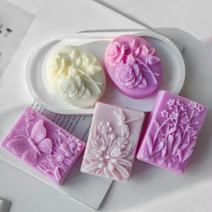 Puede incluir: Cuatro barras de jabón hechas a mano con diseños florales. Los jabones son en tonos de rosa, blanco y morado. Los jabones están dispuestos en un plato ovalado blanco. El texto "CraftMore" es visible en el plato.