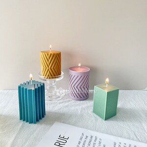 May include: Four decorative candles with different geometric patterns. The candles are in shades of yellow, purple, teal, and green. The candles are lit and sitting on a white surface.