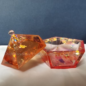 May include: A small, orange, diamond-shaped trinket box with a lid and a clear, iridescent interior. The box is decorated with gold glitter and has a small, clear knob on the lid.