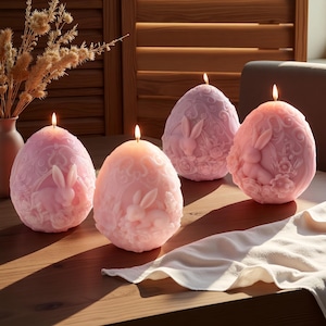 May include: Four lit, egg-shaped candles in shades of pink and purple, each with a carved bunny and floral design. The candles sit on a wooden table with a white cloth, with a vase of dried flowers in the background.