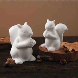 May include: Two white ceramic squirrel figurines, one standing and one sitting, each holding a nut. The squirrels have fluffy tails and are set on a dark wooden surface with acorns and a leaf.