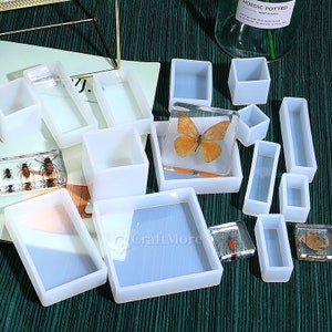 Peut inclure: Un ensemble de 12 moules en silicone transparents pour la coulée de résine. Les moules sont de formes variées, notamment des carrés, des rectangles et une forme de papillon. Les moules sont sur une surface verte.