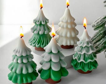 May include: A collection of Christmas tree-shaped candles with lit flames. The candles are in shades of green and white, with a brown base. The candles are arranged on a white surface, creating a festive holiday display.