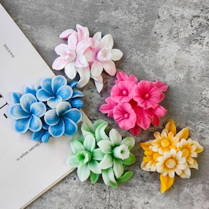 Könnte beinhalten: Fünf verschiedene künstliche Blumenarrangements in verschiedenen Farben. Die Blumen sind aus einem glatten, wachsartigen Material gefertigt und in Büscheln angeordnet. Die Blumen sind rosa, weiß, blau, grün und gelb.