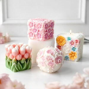 May include: A collection of decorative candles in various shapes, including a cube covered in pink flowers, a sphere adorned with pink orchids, and a square candle with colorful floral accents. One candle is lit.