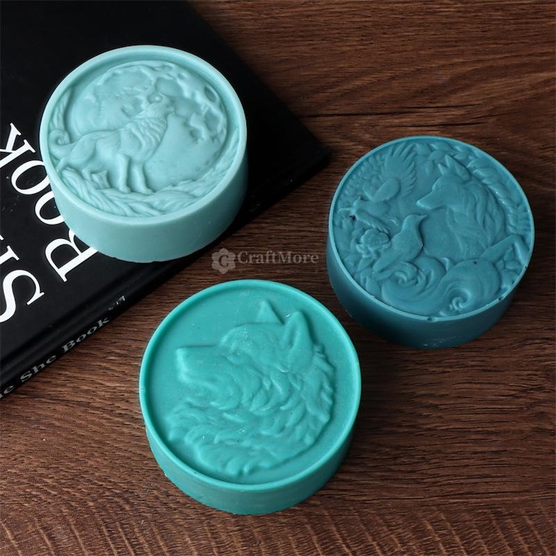 May include: Three round, teal-colored soaps with embossed animal designs. One soap features a wolf, another a wolf and moon, and the third a wolf, bird, and other elements. The soaps are on a wooden surface.