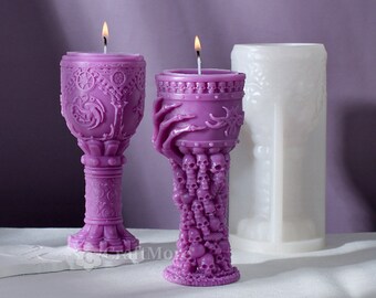 Pode incluir: Duas velas roxas acesas com designs intrincados. Uma vela tem a forma de um cálice com padrões detalhados, enquanto a outra apresenta uma mão segurando a taça da vela, sustentada por uma base de caveiras. Uma forma de vela branca no fundo.