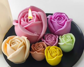 May include: A selection of rose-shaped candles in shades of pink, purple, yellow, and green, displayed on a black plate. One pink candle is lit, with a visible flame. The candles are designed to look like real roses.