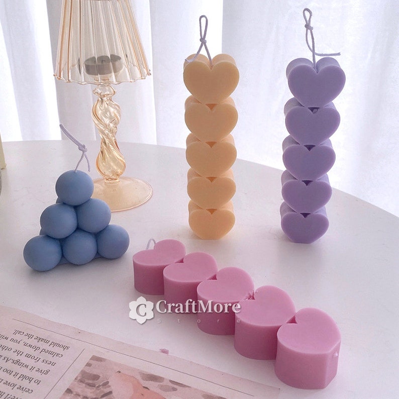 May include: Assortment of colorful heart and sphere shaped candles. The candles are arranged in a row on a white surface. The candles are made of wax and have a wick in the center. The candles are in various colors, including purple, pink, blue, and yellow.
