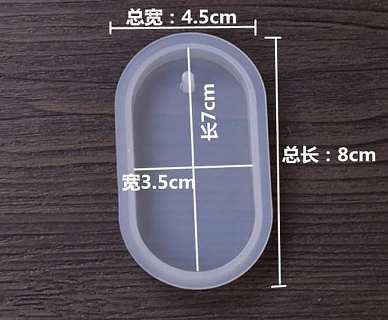May include: A clear oval-shaped silicone mold with measurements in centimeters. The mold is 8 cm long, 7 cm tall, and 3.5 cm wide. The mold is on a wooden surface.