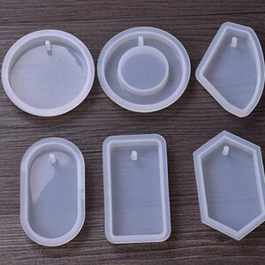 May include: Six clear silicone molds for making jewelry. The molds are in various shapes, including a circle, oval, rectangle, and a hexagon. Each mold has a small hole at the top for adding a bail or jump ring.
