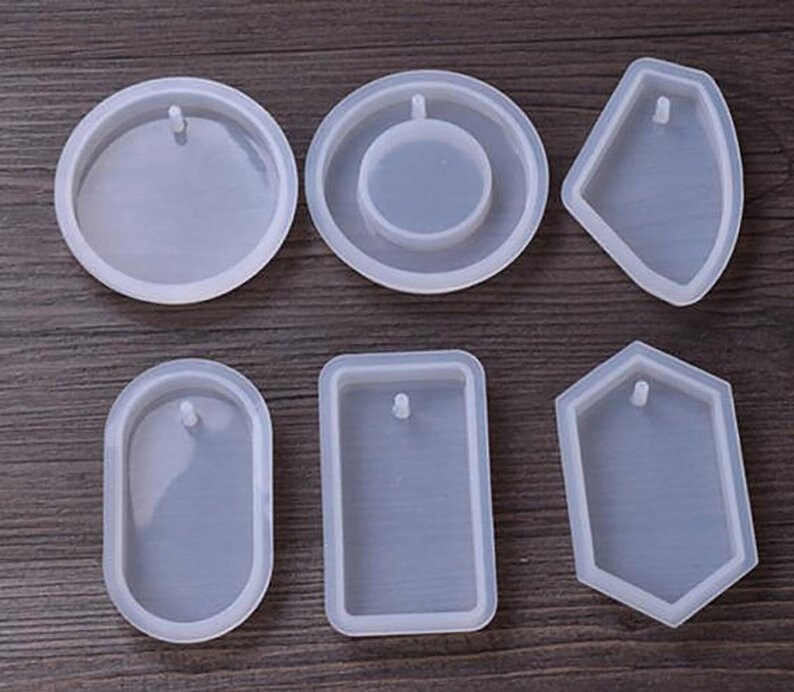 May include: Six clear silicone molds for making jewelry. The molds are in various shapes, including a circle, oval, rectangle, and a hexagon. Each mold has a small hole at the top for adding a bail or jump ring.