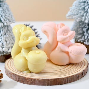 May include: Two small squirrel figurines made of soap. The squirrel on the left is yellow and holding an acorn. The squirrel on the right is pink and holding a peach.