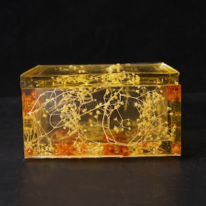 May include: A rectangular, clear resin box with a yellow tint, encasing dried flowers and foliage. The box has a lid and is set against a dark background. The flowers are a light yellow colour.