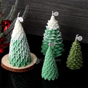 May include: Four green and white Christmas tree candles of varying sizes. The candles are decorated with green leaves and have a white top. The candles are sitting on a wooden surface.