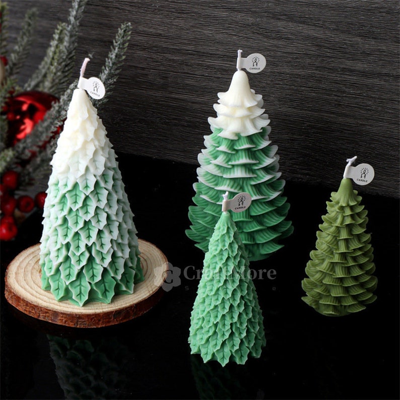 May include: Four green and white Christmas tree candles of varying sizes. The candles are decorated with green leaves and have a white top. The candles are sitting on a wooden surface.