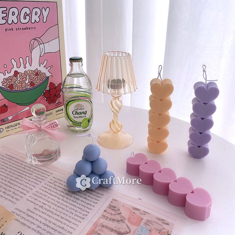 May include: Assortment of colorful heart-shaped and round candles. The candles are arranged on a white surface. The candles are in various shades of pink, purple, and blue.