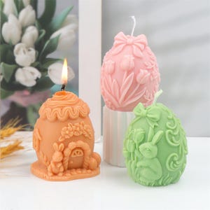 Könnte beinhalten: Drei Ostereier-Kerzen. Eine ist angezündet, orange und zeigt ein Kaninchen und ein Haus. Die anderen beiden sind rosa und grün, verziert mit Schleifen, Blumen und einem Hasen. Diese dekorativen Kerzen sind perfekt für Ostern.