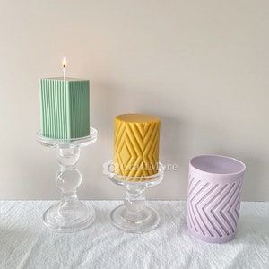 May include: Three candles on glass candle holders. The candles are different shapes and sizes. One candle is green and has vertical lines, one is yellow and has a diamond pattern, and one is purple and has a chevron pattern.