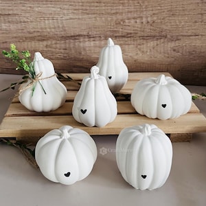 Könnte beinhalten: Sechs weiße Keramik-Kürbisse mit schwarzen Herz-Akzenten. Die Kürbisse sind auf einer Holzoberfläche angeordnet.