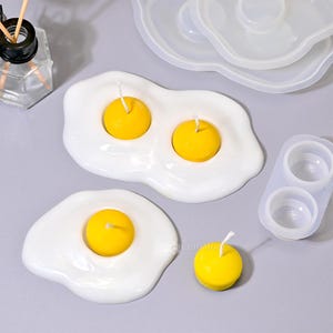May include: Three silicone moulds for making candles in the shape of fried eggs. The moulds are white and have a realistic egg shape. The moulds are shown with yellow candles in them.