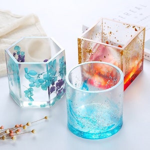 Puede incluir: Tres recipientes de resina transparente con diferentes formas y diseños. El primer recipiente es de forma hexagonal con flores azules y moradas prensadas. El segundo recipiente es de forma cuadrada con motas doradas y una resina rosa y naranja en su interior. El tercer recipiente es de forma redonda con una resina azul en su interior y purpurina plateada.