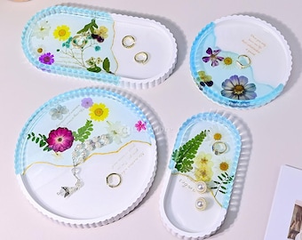 May include: Four resin jewelry trays with dried flowers and gold accents. The trays are oval and round, with a white base and a light blue border. Some trays have rings and a necklace.