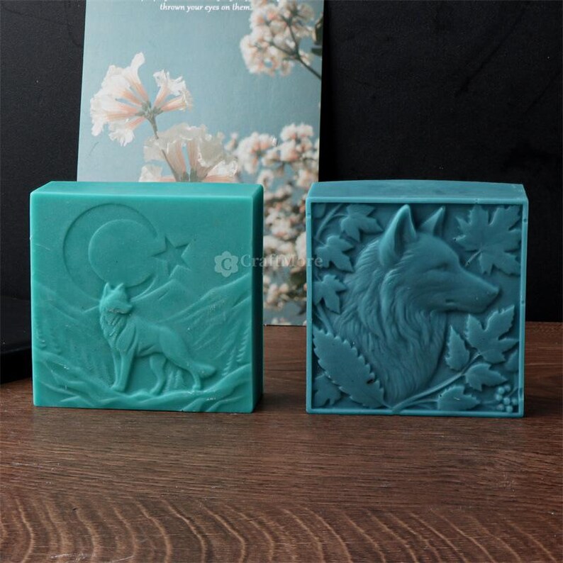 May include: Two teal-colored, square soaps. One soap features a wolf with a crescent moon and star design. The other soap has a detailed wolf head surrounded by leaves. Both soaps have a carved relief design.