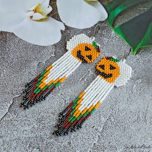 Op de afbeelding: Een paar kralen Halloween-oorbellen. Elke oorbel heeft een lachend pompoengezicht in oranje en zwart, met een witte kralenwolk erboven. De oorbellen hebben een lange franje van witte, oranje, groene en zwarte kralen.