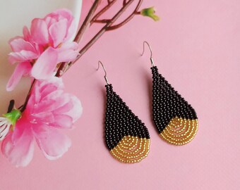 May include: A pair of teardrop-shaped earrings with black and gold beads. The earrings are made of black beads with a gold bead accent at the bottom.