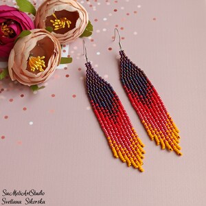 May include: A pair of long, beaded earrings with a gradient of purple, red, and yellow beads. The earrings are hanging from silver hooks.
