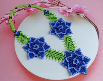 May include: A beaded necklace featuring three blue flower designs and green leaf accents. The necklace is displayed on a white plate.